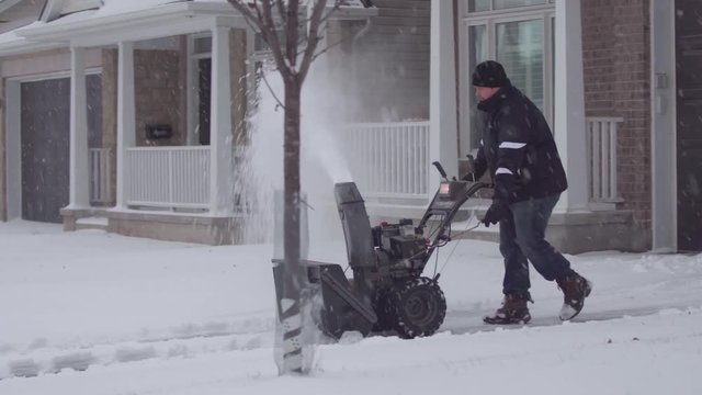 A wide-shot of a man using a snow-blower on his driveway in the suburbs.