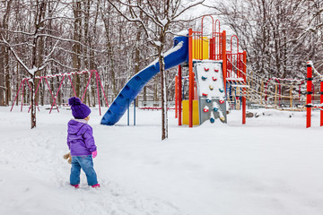 Little girl walking outside. Kid looking at the snowy children playground. Snow winter.