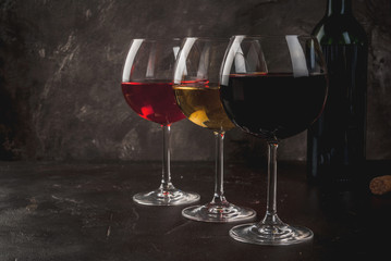 Selection of three kinds (Varieties) of wine - red, white and pink. In glasses, with an open bottle. On a dark concrete background. Copy space