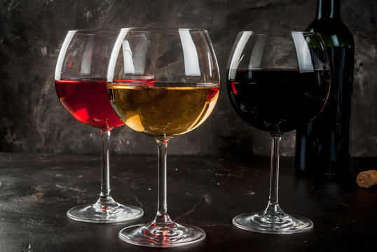 Selection of three kinds (Varieties) of wine - red, white and pink. In glasses, with an open bottle. On a dark concrete background. Copy space