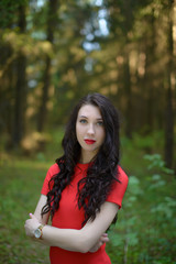 A beautiful model girl dressed in a red dress is standing in a mystical forest alone