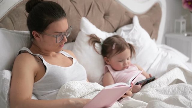 Family Day Off at the Bedroom. Clever Baby Girl is Using Smartphone. Brunette Woman in Eyesglasses is Reading a Book Lying on White Bed.