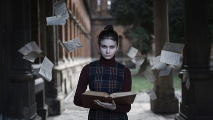 A girl with a book and a cold look. Around her pages flew out of the book against the backdrop of the old university. Concept: stopping time.