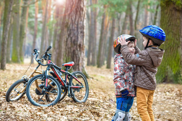 Obraz premium Two little kids boys, best friends in autumn forest. Older brother helping younger child to put his bike helmet. Happy siblings with bicycles.