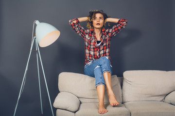 beautiful young woman sitting on couch against grey wall,curly hair,plaid shirt,blue jeans,home...