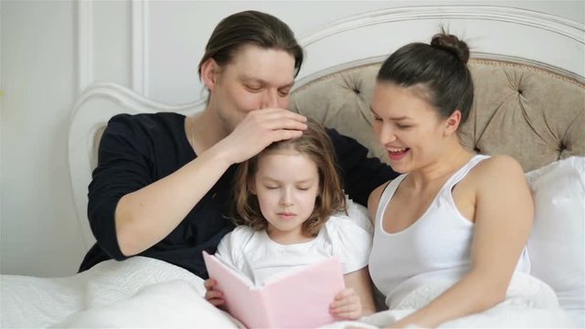 Beautiful Little Girl is Reading Aloud the Book Lying on the Bed with Her Parents. Handsome Father is Kissing His Clever Child.