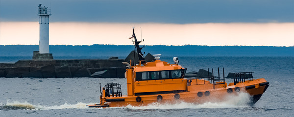 Orange pilot ship moving at speed past the lighthouse in Riga