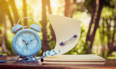 Blue alarm clock and notebook on a wooden desk with blurred nature background.