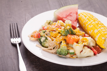 Vegetable Primavera with white sauce and shredded cheese with corn on the cob and slice of watermelon distant horizontal shot