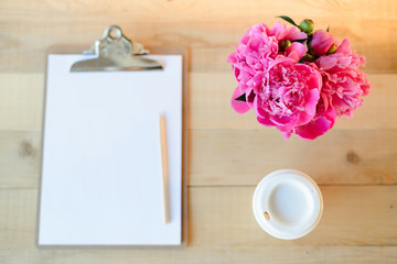 Top view flat lay of blank clipboard with pencil, cup of coffee and pink peony bouquet on wooden background