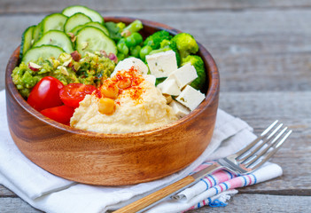 Bowl of bright healthy vegan lunch: vegetable salad with tofu, hummus and guacamole. Buddha rainbow wooden bowl, vegan concept.