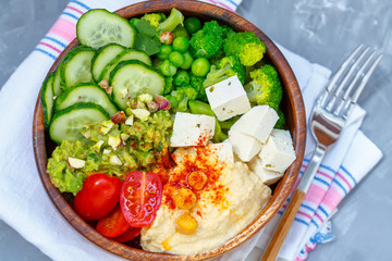 Bowl of bright healthy vegan lunch: vegetable salad with tofu, hummus and guacamole. Buddha rainbow wooden bowl, vegan concept.