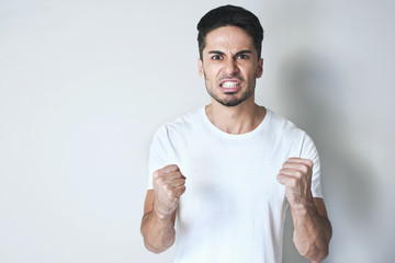 Young man is fighting. Serious man is angry and wants second try, standing in a white loose t-shirt with hands in fists against grey background