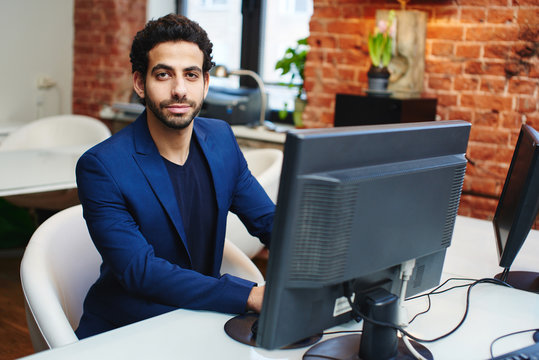 An Arab businessman in a jacket sitting on workplace behind computer in office