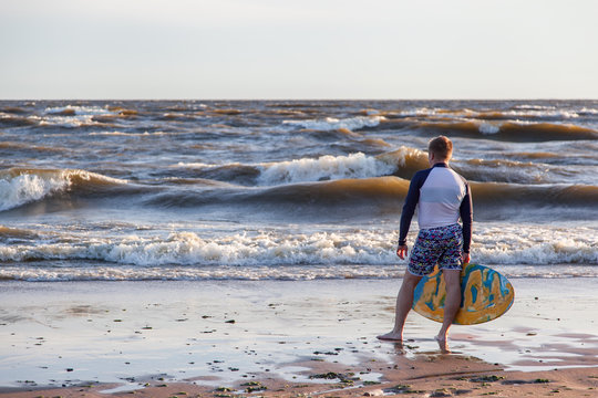Young sportive man is running after his yellow and blue skimboard on the beach on a sunny day and on the sky there are dark clouds