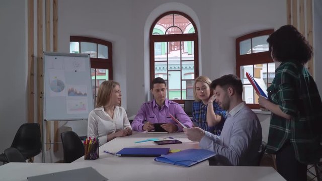 Young casual group of people sitting in the boardroom. Brunette giving folders with presentation or report coworkers. Professional men and women in stratup company reading paper documents with
