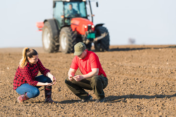 Young farmers examing  dirt while tractor is plowing fields