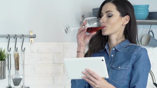 Young woman watching movie on tablet computer and drinking wine in kitchen at home
