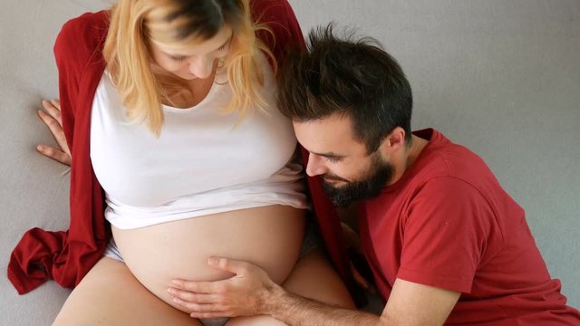 Happy bearded man touching pregnant woman belly, young excited couple expecting baby relaxing together on the bed at home.