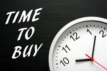 The phrase Time to Buy written on a blackboard next to a modern wall clock as a reminder for when to invest