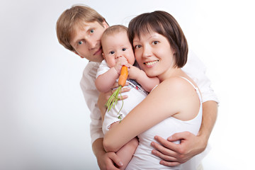 Mom and Dad are hugging a baby who is eating a carrot. Happy family on a clean white background