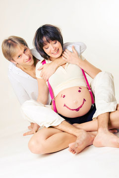 Husband hugging his pregnant wife sitting on the floor. Woman's belly is painted with a smiley