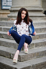 Young stylish brunette girl on shirt, pants, jeans jacket and high heels shoes, posed at stone stairs. Street fashion model concept.