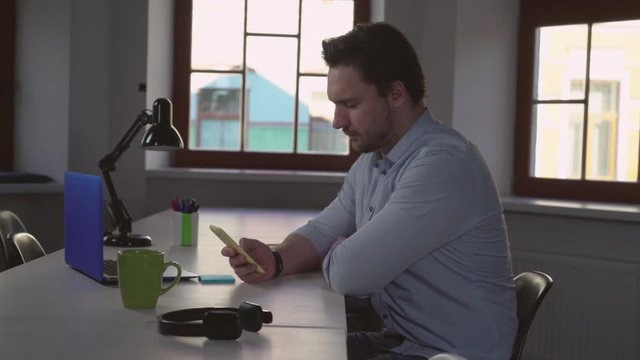 Portrait adult businessman sitting at the table in modern office. Handsome professional man holding smartphone using app. Male wearing in casual shirt alone in room with windows.