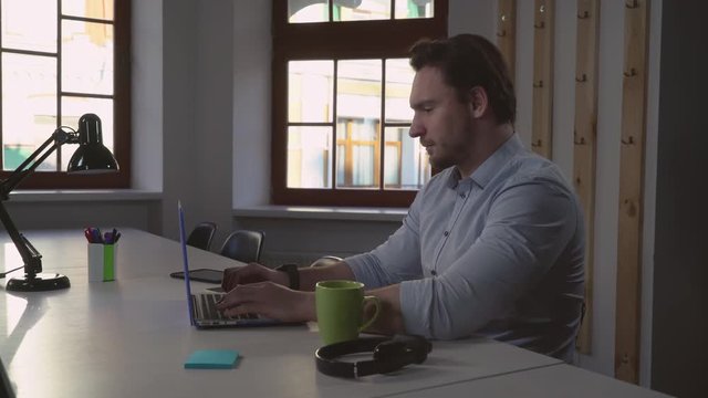 portrait man computing in office. Handsome caucasian worker sitting at the desk with table-lamp focused on screen laptop. Professional businessman concentrated typing in empty room