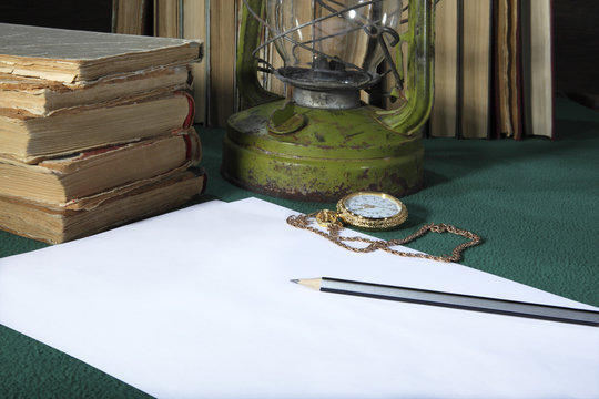 On the desktop are: a clean white sheet of paper, a simple pencil, old books, pocket watch on a gold chain and a kerosene lamp. Retro stylized photo