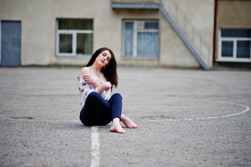 Fototapeta na wymiar Young stylish teenage brunette girl on shirt, pants and high heels shoes, sitting on pavement and posed background school backyard. Street fashion model concept.