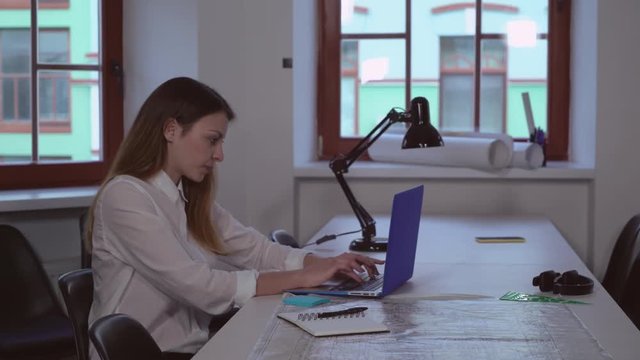 Young caucasian architect working in creative office. Professional designer sitting the the table with blueprint. Lady wearing in casual dress typing on computer.