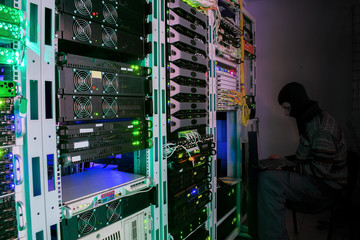 The hacker in a mask with the laptop sits at a chair in datacenter server room