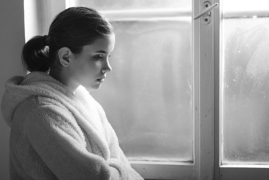 Beautiful ill sad girl, cancer patient in night gown looking through hospital window