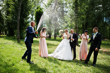 Naklejka na ściany i meble Fashionable wedding couple with bridesmaids and best mans with explosion of champagne bottle at forest on sunny wedding day.