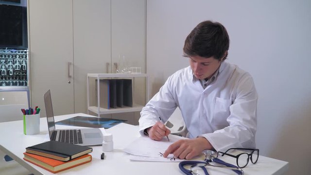 Caucasian doctor with friendly smile giving X-ray of the lungs patient. Professional man sitting in the office wearing in white coat. On the desk laptop, prescription and bottle with medicine