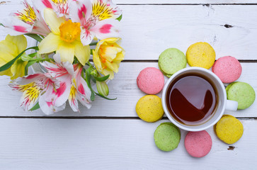Multicolored macaroons, flowers and cup of hot tea on a wooden table.