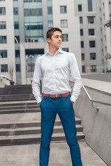 Portrait of cheerful businessman standing outdoors near the business center. Young man holds hands in his trouser pockets.