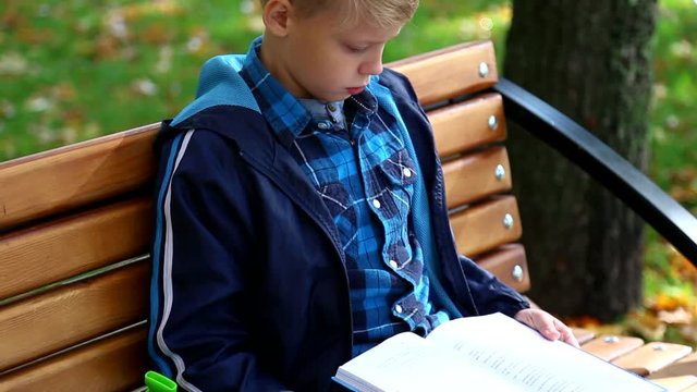 9 years old boy sits on wooden bench in city park after school and reads  favorite book. Real time full hd video footage.