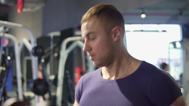 A young man is practicing at the gym. He pumps his arm muscles with dumbbells. 4K