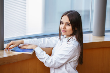 Portrait of smiling Caucasian brunette young beautiful girl woman student with long dark hair and brown eyes in white shirt holding exercise book standing by window in college university