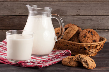 jug and glass of milk with oatmeal cookies in a wicker basket on a wooden background