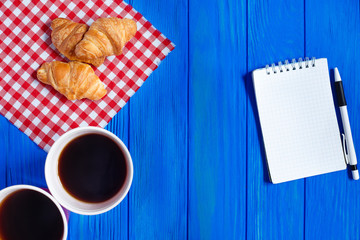 Two cups of coffee, croissants and open notepad with pen on blue wooden table