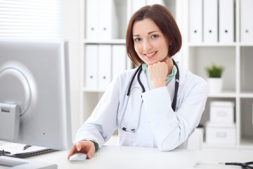 Young brunette female doctor sitting at a desk and working on the computer at the hospital office.  Health care, insurance and help concept. Physician ready to examine patient