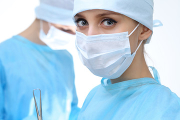 Close up of surgeon woman looking at camera while colleagues performing in background in operation room, isolated. Surgery and medicine concept