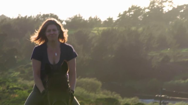 4k Cine long lens of adult equestrian woman riding English style saddle on a Thoroughbred horse trotting toward camera and smiling with sun hitting her back hair as she gets full frame SDF1