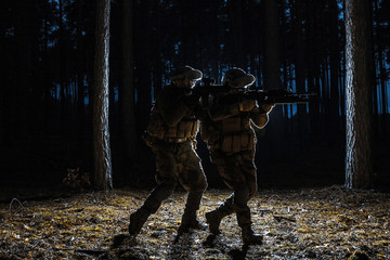Black silhouettes of pair of soldiers in the forest moving in battle operation. Back light