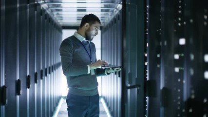 Male IT Technician Working on a Laptop Standing Before Open Server Rack Cabinet in Big Dara Center. Shot on RED EPIC-W 8K Helium Cinema Camera.