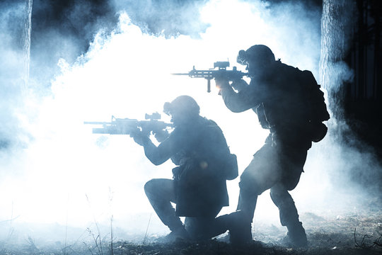 Black silhouettes of pair of soldiers in the smoke moving in battle operation. Back light