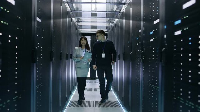 Caucasian Male and Asian Female IT Technicians Walking in Data Center with Rows of Rack Servers. They Have Discussion, She Holds Tablet Computer. Shot on RED EPIC-W 8K Helium Cinema Camera.
